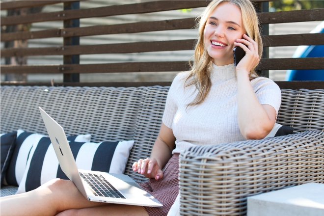 Woman sitting outside on a couch as she is smiling and looking at a laptop.